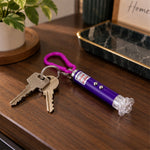 A purple laser pointer keychain with a pink clip rests on a wooden surface next to a pair of silver keys.