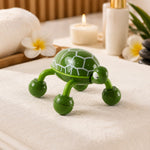 A green turtle-shaped massager with white shell patterns sits on a white towel next to a lit candle and spa items.