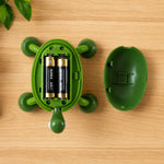 The green turtle massager is open, revealing two AA batteries inside, resting on a wooden surface next to its shell.