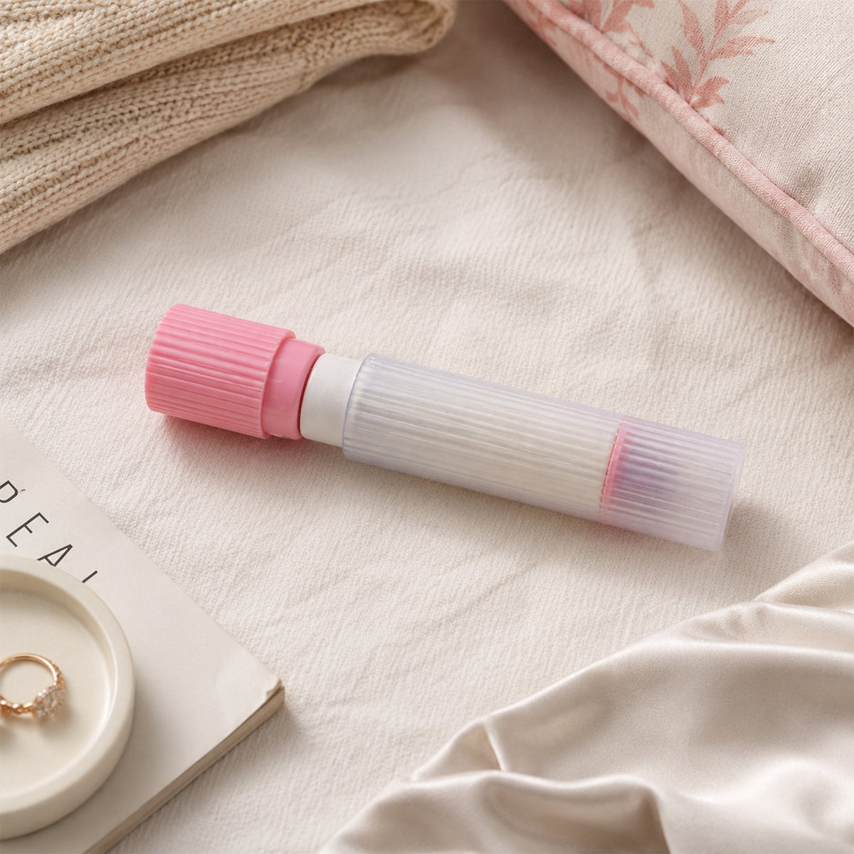 A pink and white lint roller rests on a white textured surface next to a ring on a dish and a folded knit blanket.