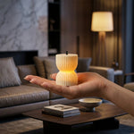 A hand holds a single glowing, ribbed, hourglass-shaped LED candle above a dark coffee table in a cozy living room.