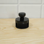 The black detailing brush is placed inside its cylindrical base on a light wooden surface, with a white tiled wall behind.