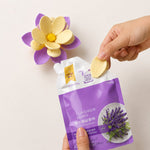 A hand inserting a yellow petal into a purple and yellow flower clip, next to a lavender bead pouch.