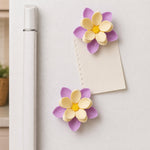 Two purple and yellow flower clips holding a small note to a white refrigerator door.