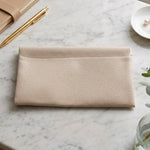 A cream textured faux leather pouch lying flat on a white marble surface, next to a gold pen and a small white dish with
