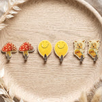 Six assorted cartoon wall hooks: two red mushrooms, two yellow smiling faces, and two sheep on skateboards, on a wooden tray.