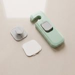A disassembled green and grey child safety lock with two white adhesive pads on a light-colored surface.