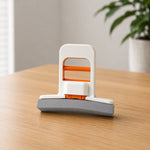 A hand uses a white and orange mini sponge mop to clean up a spilled coffee stain on a white desk next to a laptop.