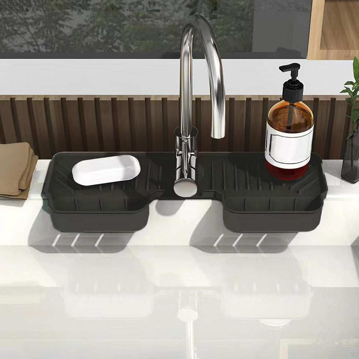 A black silicone faucet splash guard with a bar of soap and a pump bottle on a white kitchen sink, with a window and wooden