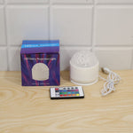 A white LED projection light, its box, and a remote control on a light wooden surface against a white tiled wall.