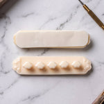 Two beige self-adhesive cable organizer clips on a white marble surface; one shows the adhesive backing, the other shows the