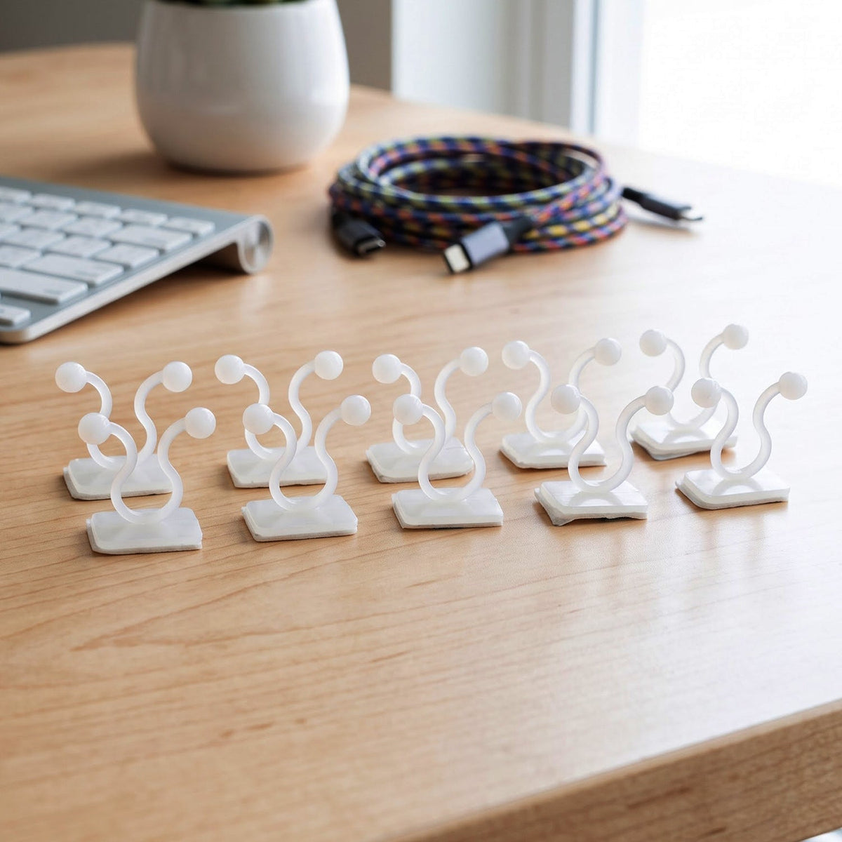 Eight white plastic self-adhesive plant clips stand upright on a light wooden desk next to a keyboard and coiled cables.