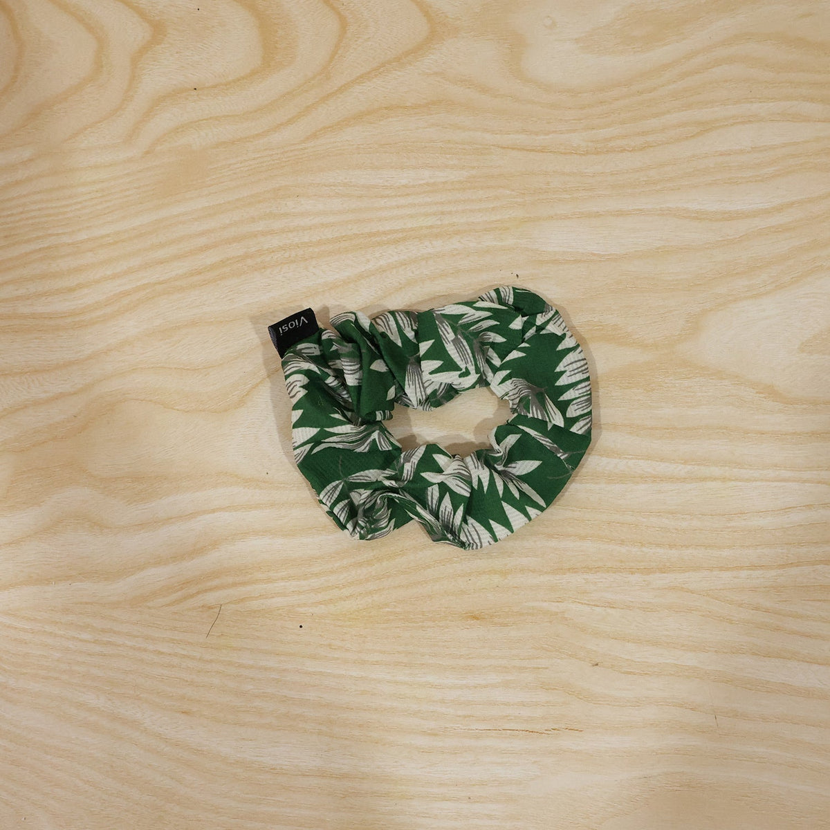 A green and white leaf print scrunchie with a black 'viosi' tag is placed on a light wooden surface.