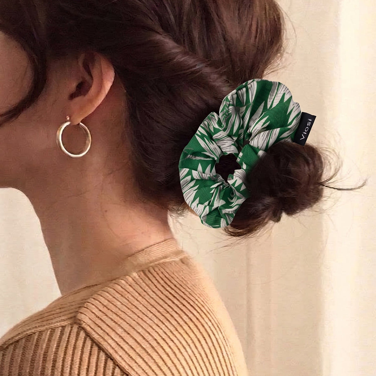 A person with brown hair wears a green and white leaf print scrunchie in a bun, with a gold hoop earring visible.