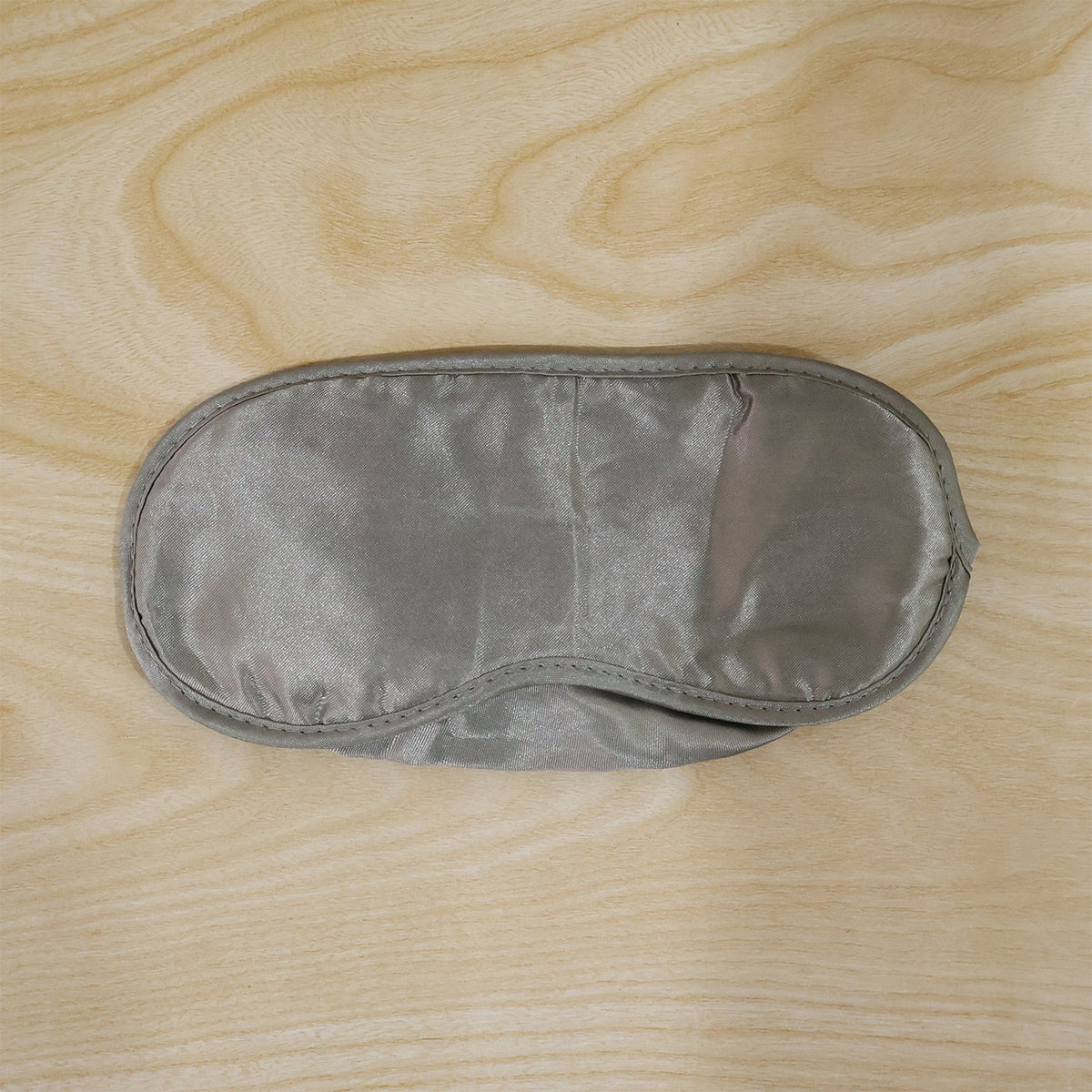 A grey satin sleeping eye mask with a smooth surface is laid flat on a light wooden background.