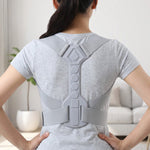 Back view of a person wearing a light gray posture corrector, standing with hands on hips in a bright room.
