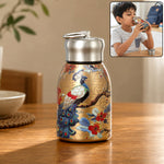Golden stainless steel vacuum bottle with a peacock and floral print on a wooden table, with a small inset of a boy drinking