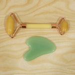 A yellow facial roller with rose gold accents and a green gua sha tool are positioned on a light wooden surface.