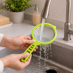 Hands hold a green plastic strainer with stainless steel mesh under running water in a kitchen sink.
