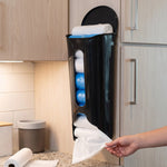 A hand pulls a white plastic bag from a black wall-mounted dispenser, filled with white and blue bags.