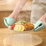 Two hands wearing green silicone mini oven mitts hold a steaming plate of spaghetti with lemon and tomatoes.