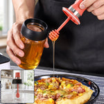 A person drizzles honey from a glass dispenser with a red silicone dipper onto a pizza slice.