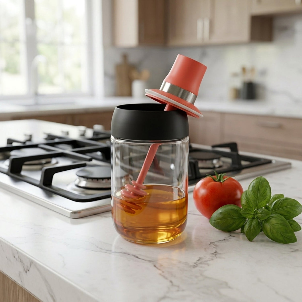 A glass honey dispenser with a red dipper and black lid sits on a kitchen counter next to a tomato and basil.