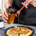 A person drizzles honey from a glass dispenser with a red silicone dipper onto a pizza slice. – view 8