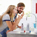 A couple brushes their teeth in a bathroom, with two tube squeezers holding toothpaste tubes on the counter.