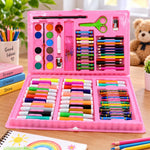 An open pink art kit displays watercolors, crayons, colored pencils, markers, scissors, and a ruler on a wooden table.
