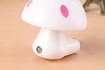 A close-up of a white mushroom-shaped night light with pink spots, showing the light sensor and plug on a textured surface.
