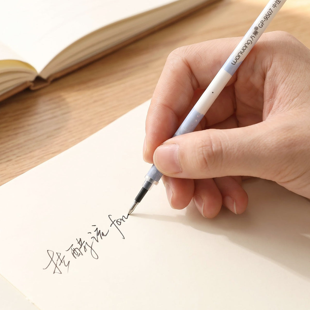 A hand writes in a foreign language on a light-colored paper with a white and blue pen refill, with a notebook.
