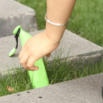 A hand reaches for a green banana-shaped umbrella case resting in green grass between concrete slabs.
