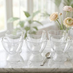Six clear plastic ice cream bowls with a small silver spoon on a white marble surface.