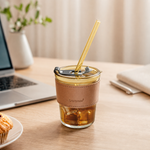 An amber glass cup with a brown leather sleeve and a straw sits on a desk with a laptop and a muffin.
