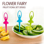 Three colorful dancing doll fruit forks, pink, blue, and green, on a wooden board with sliced kiwi and pineapple.