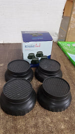 Four black round washing machine support legs are displayed in front of a blue and white product box on a wooden surface.