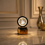 A golden base lamp with a round crystal displaying a glowing Ganesha, on a marble table with a golden vase.