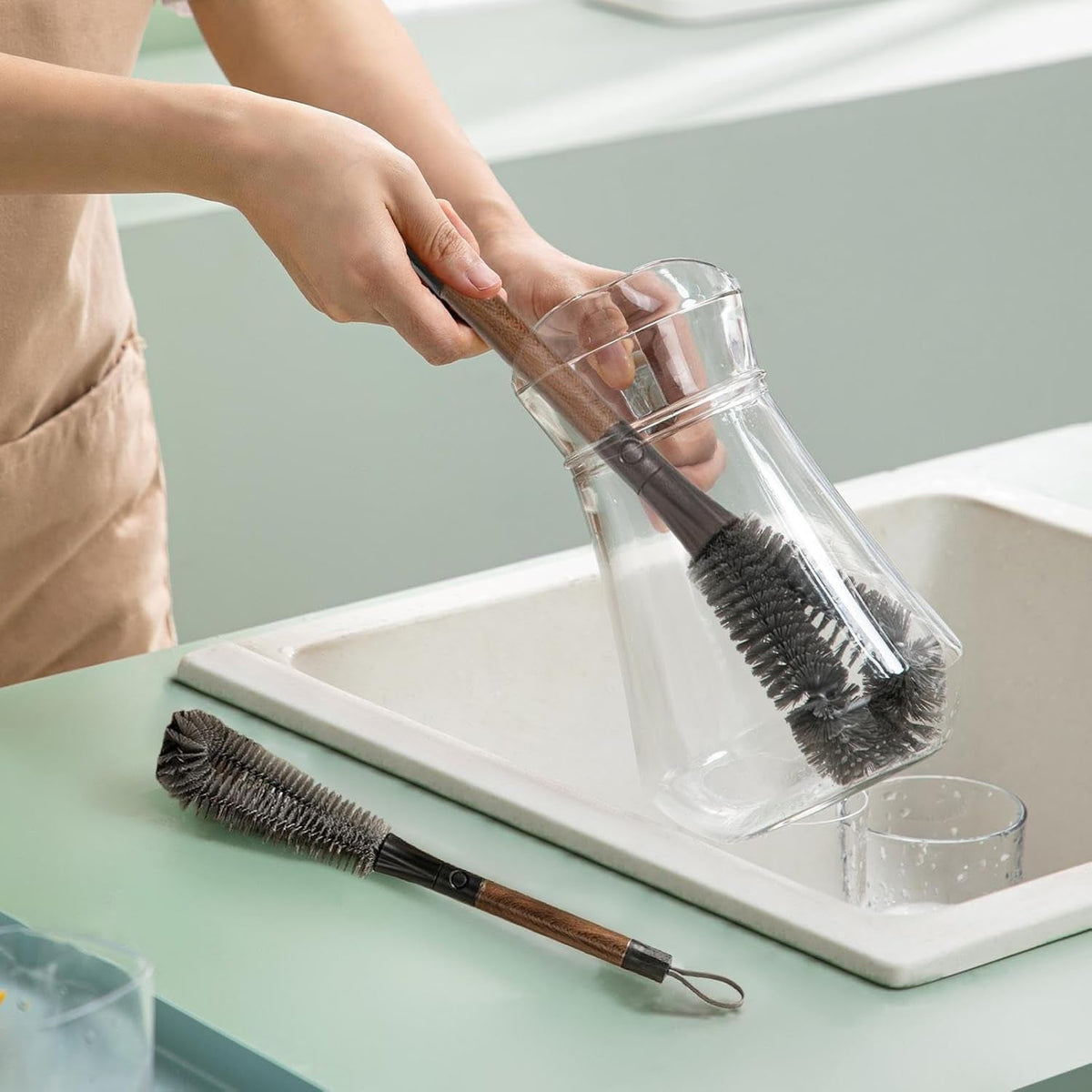 A person cleans a clear glass carafe with a long-handled grey brush, with another brush resting on the counter.