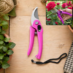 A pink garden pruner with a black wrist strap lies on a wooden surface next to potted plants and twine.