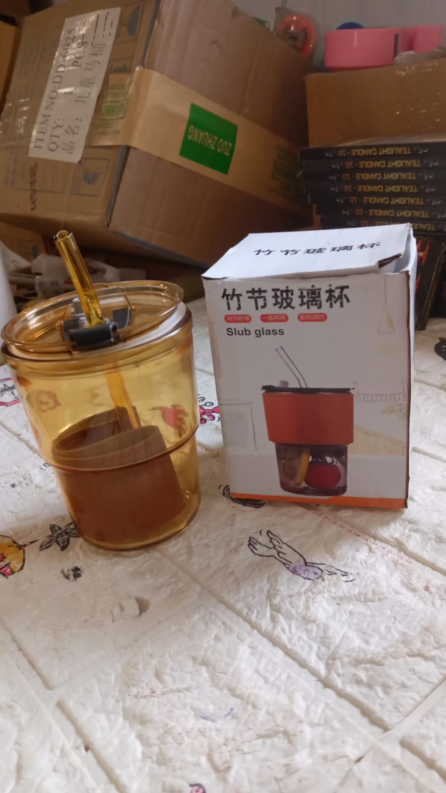An amber glass cup with a straw and a brown sleeve sits next to its product box on a patterned surface amidst cardboard