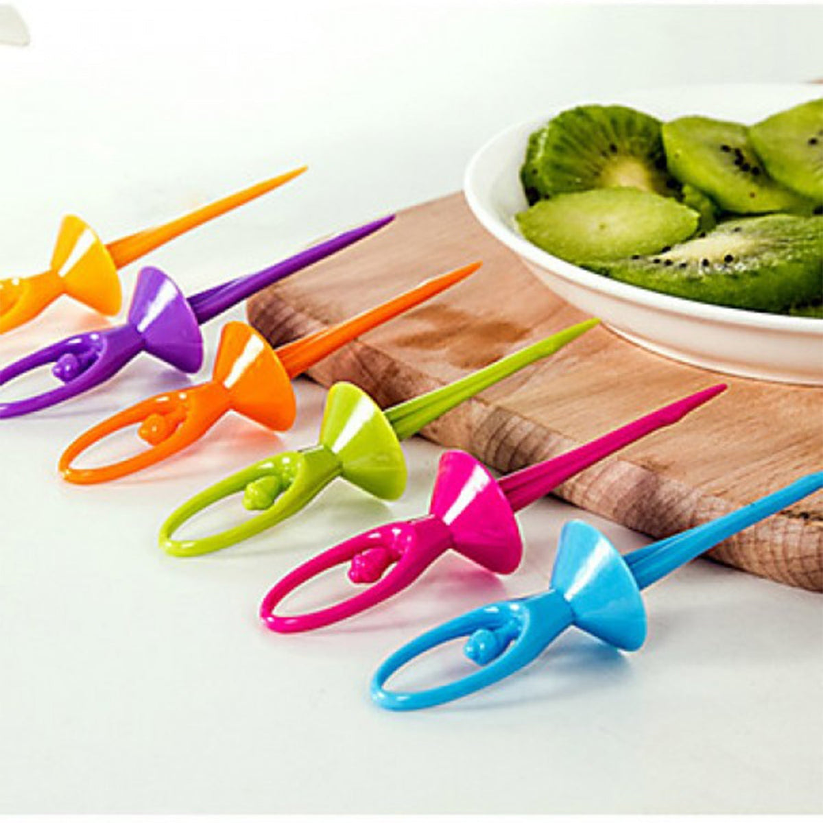 Six colorful plastic dancing doll fruit forks on a wooden board with a bowl of sliced kiwi.
