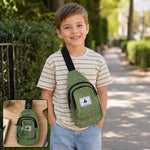 A smiling boy wears an olive green canvas sling bag with a black strap over his shoulder, walking outdoors.