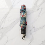 A folded floral umbrella, secured with a dark green strap, rests on a white marble surface, showing its black handle and