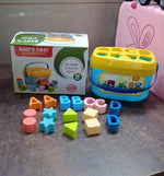 A blue and yellow shape sorter bucket and colorful blocks are displayed next to their product box, which reads "BABY'S FIRST