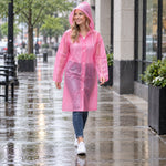 A smiling woman in a pink translucent rain coat with a hood walks on a wet city sidewalk.