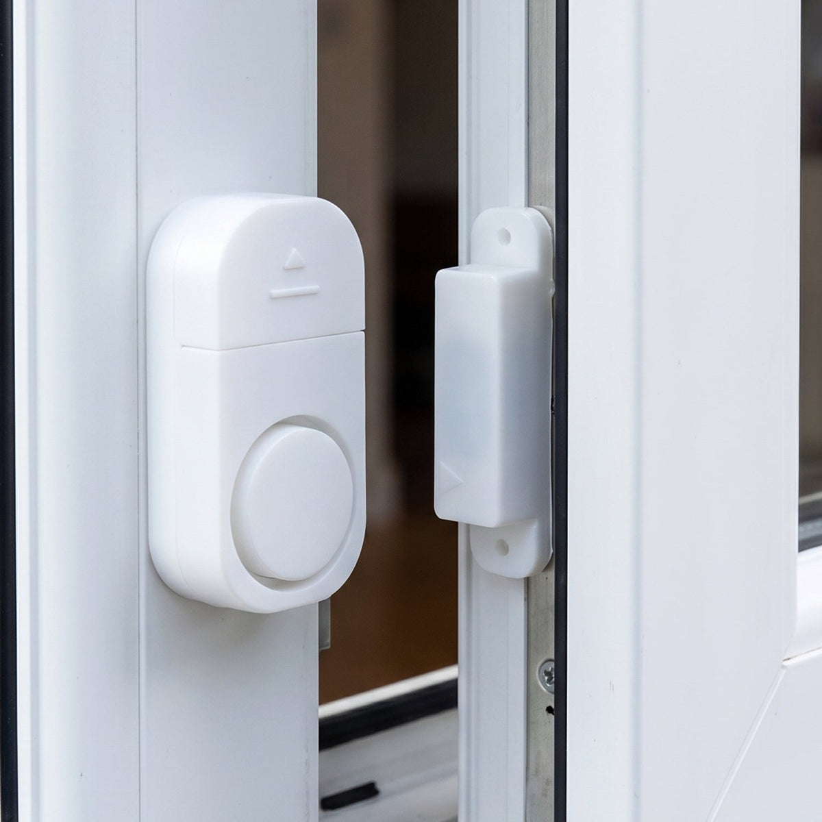 A white plastic magnetic alarm is attached to a white window frame, with its smaller magnetic component on the adjacent
