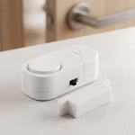 The white magnetic door alarm and its smaller magnetic piece are on a white surface, with a blurred wooden door in the