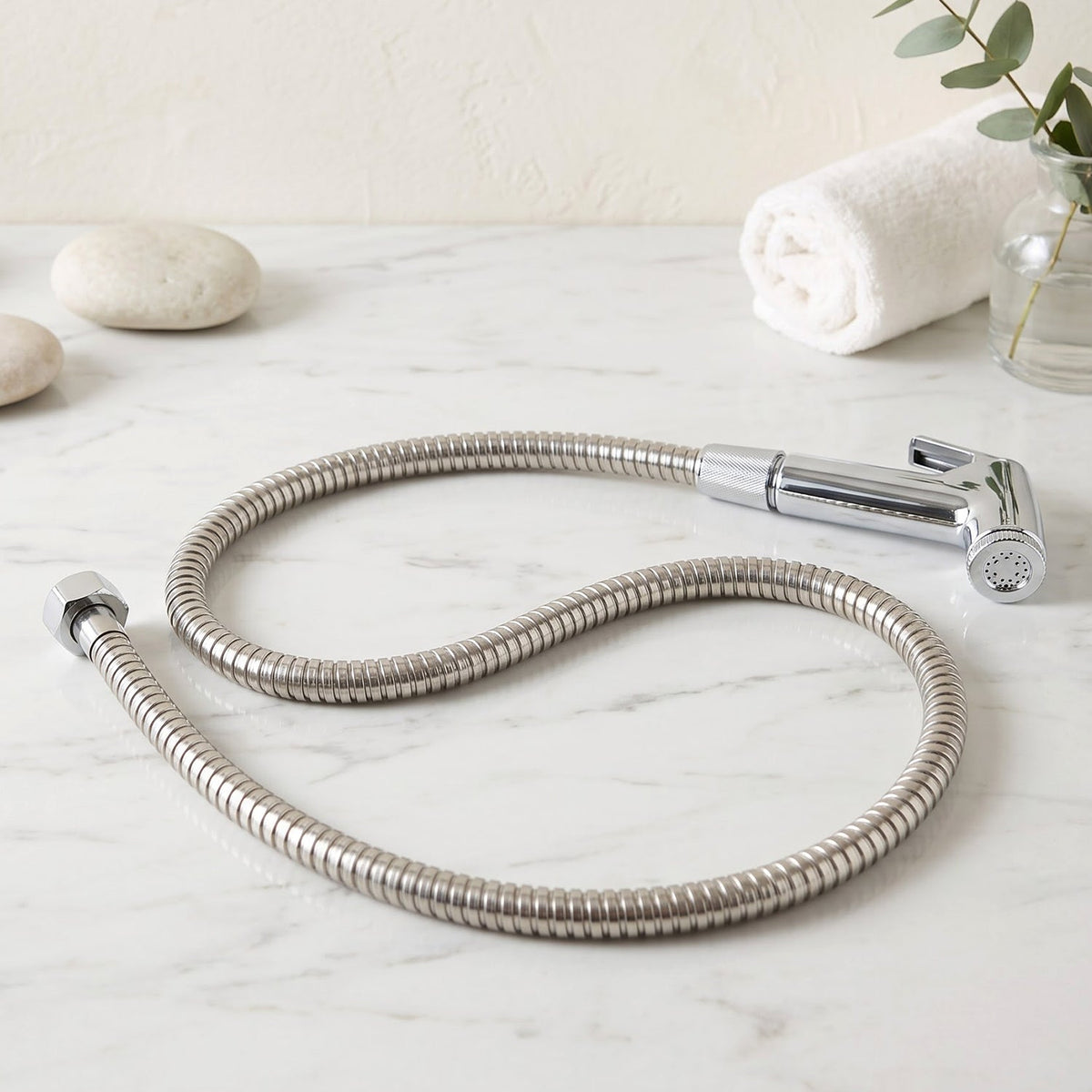 A coiled stainless steel hose with a chrome bidet sprayer lies on a white marble surface with decorative stones.