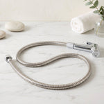 A coiled stainless steel hose with a chrome bidet sprayer lies on a white marble surface with decorative stones.
